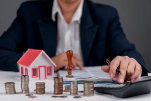 Businessman calculating real estate investment, with house model, coins, and calculator.