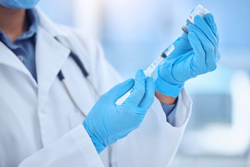 A close-up shot of a doctor or medical professional in a white lab coat and blue gloves, holding a syringe and a small glass vial.